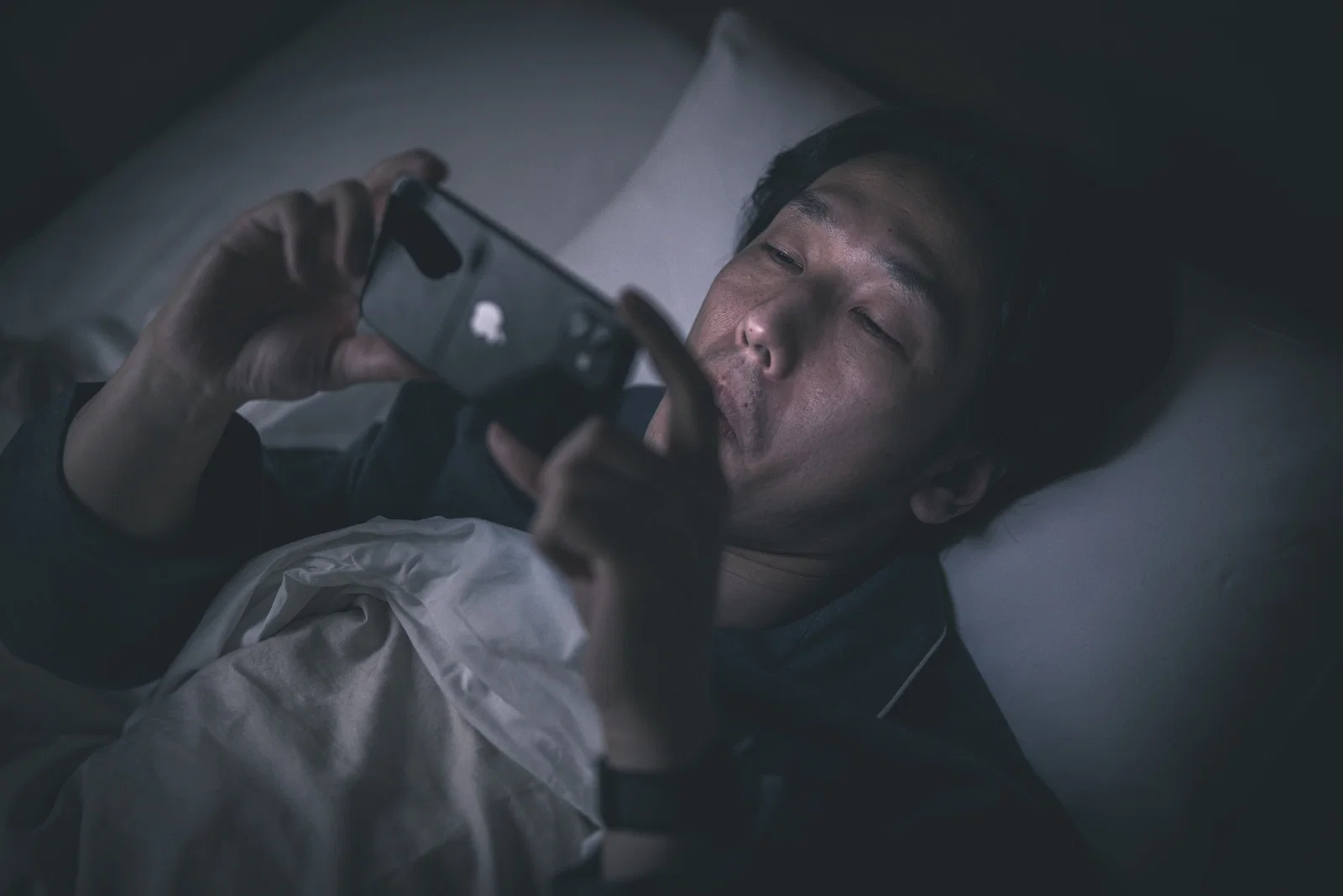 室内のベッドで横になった男性一人が、暗がりでスマートフォンを操作している