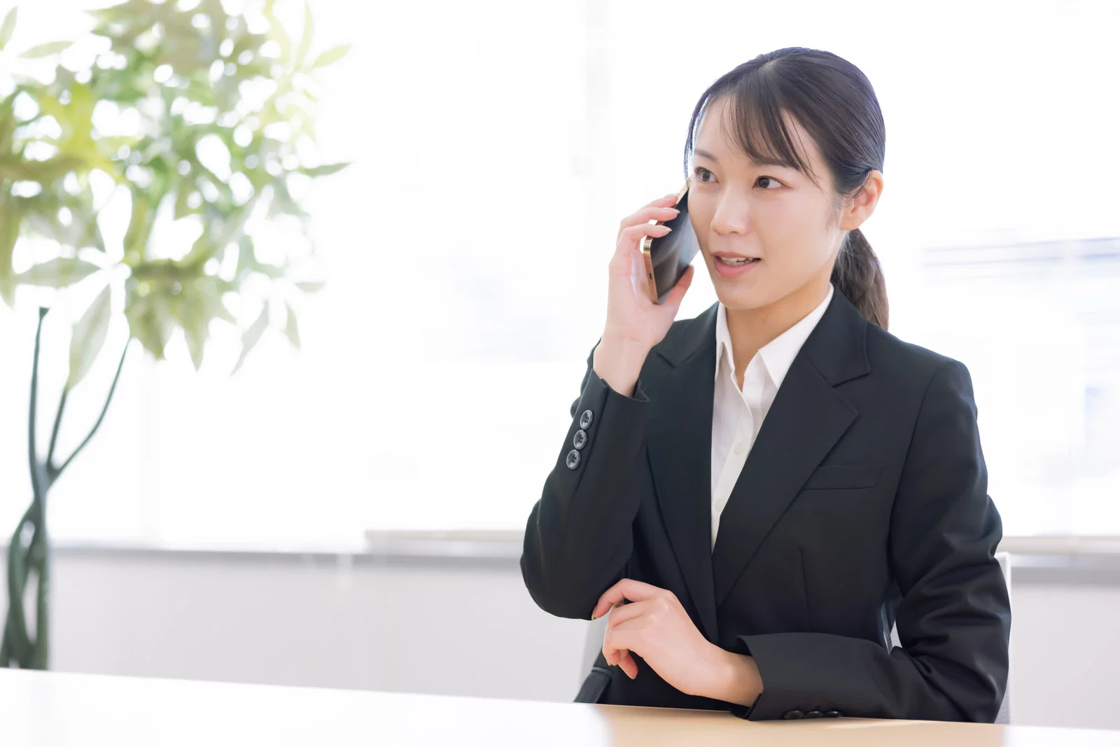 明るいオフィスの窓際で机に向かいスマートフォンで通話するスーツ姿の女性1人。
