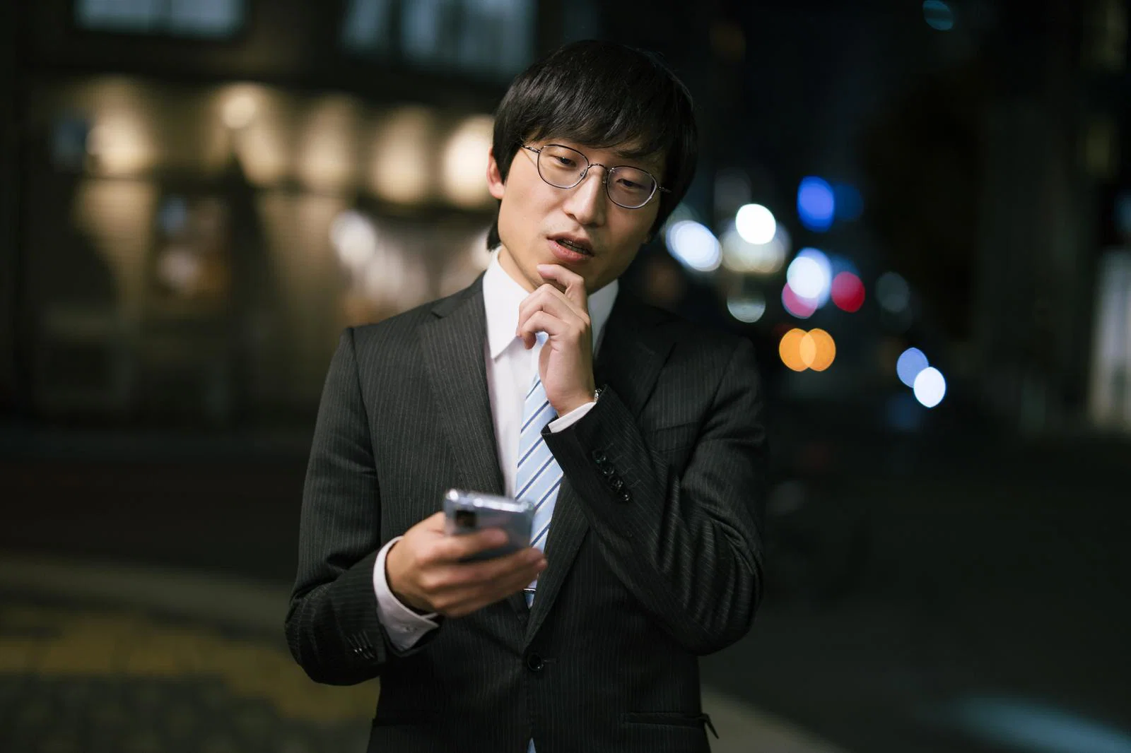 夜の屋外で、スーツ姿の男性1人がスマートフォンを見ながら考え込んでいる
