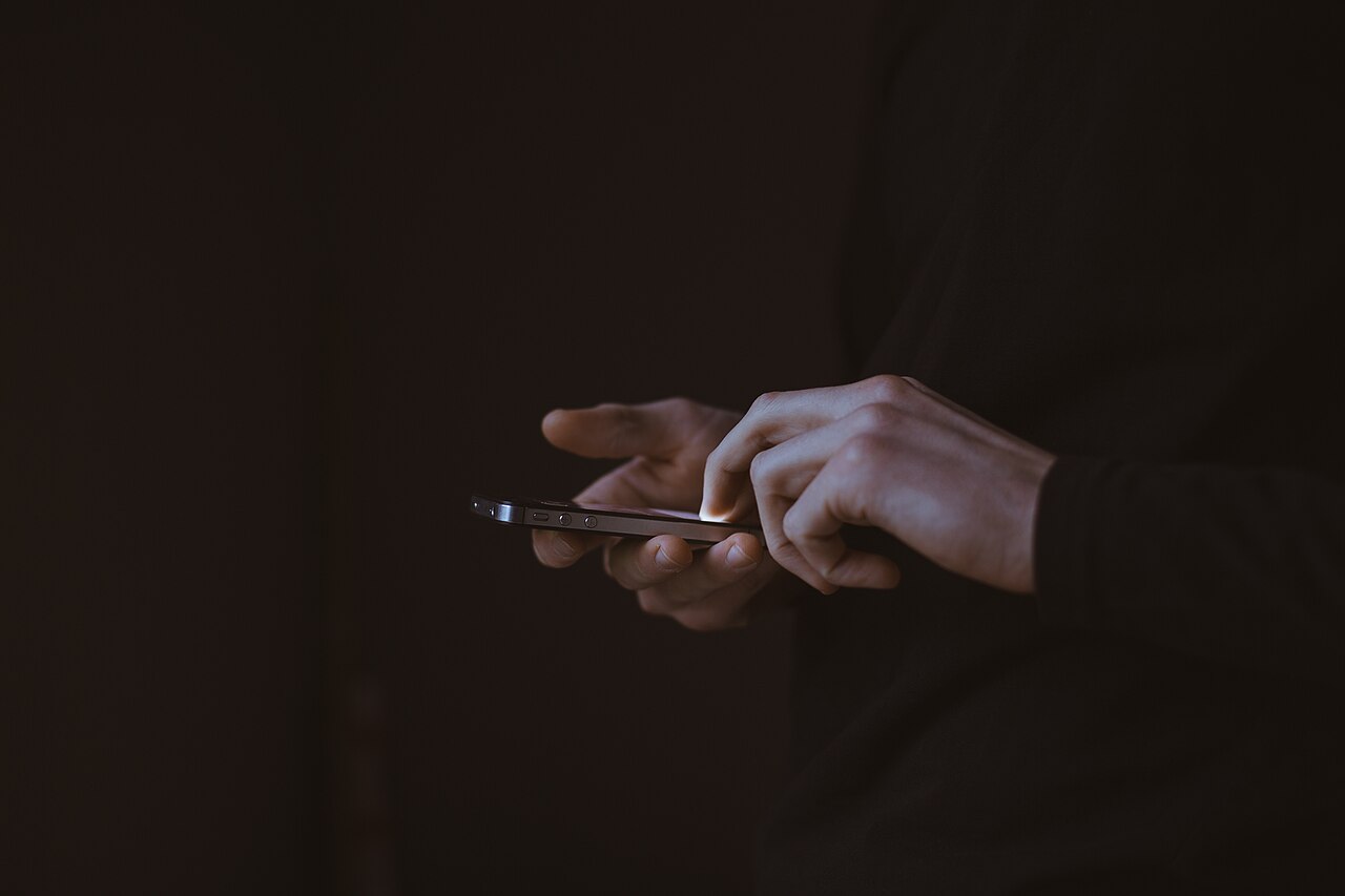 暗い室内で黒い服の人物がスマートフォンを両手で操作している接写。
