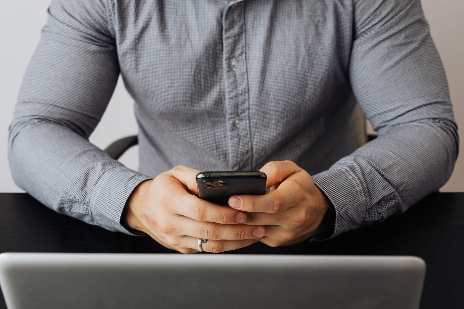 デスクに座る男性がノートPCの前で黒いスマートフォンを両手で操作している