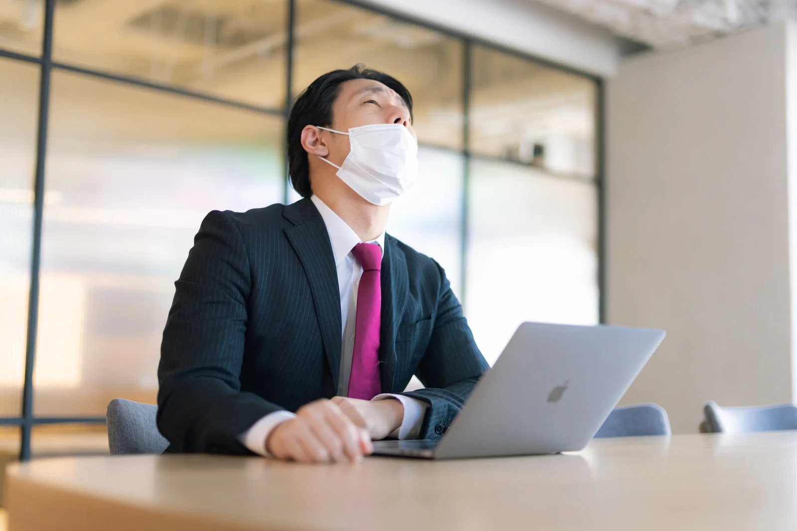 屋内会議室で、マスクをしたスーツ姿の男性1人が天井を見上げノートPCの前に座っている