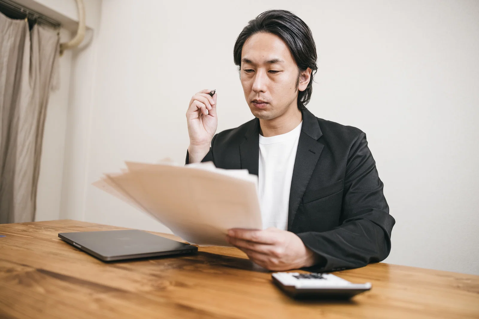 室内の木製テーブルで、スーツ姿の男性一人が書類とノートパソコンを前に読み込む様子