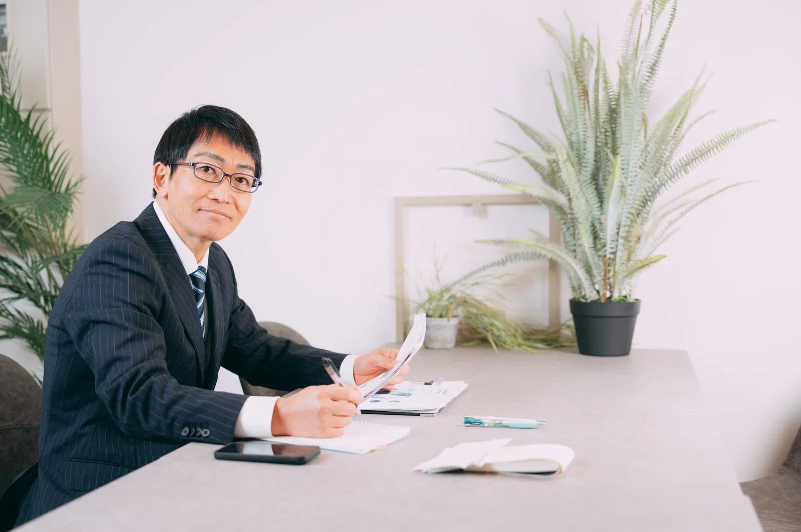 観葉植物のある室内でスーツの男性一人が机に向かい書類にペンを走らせながらこちらを見ている
