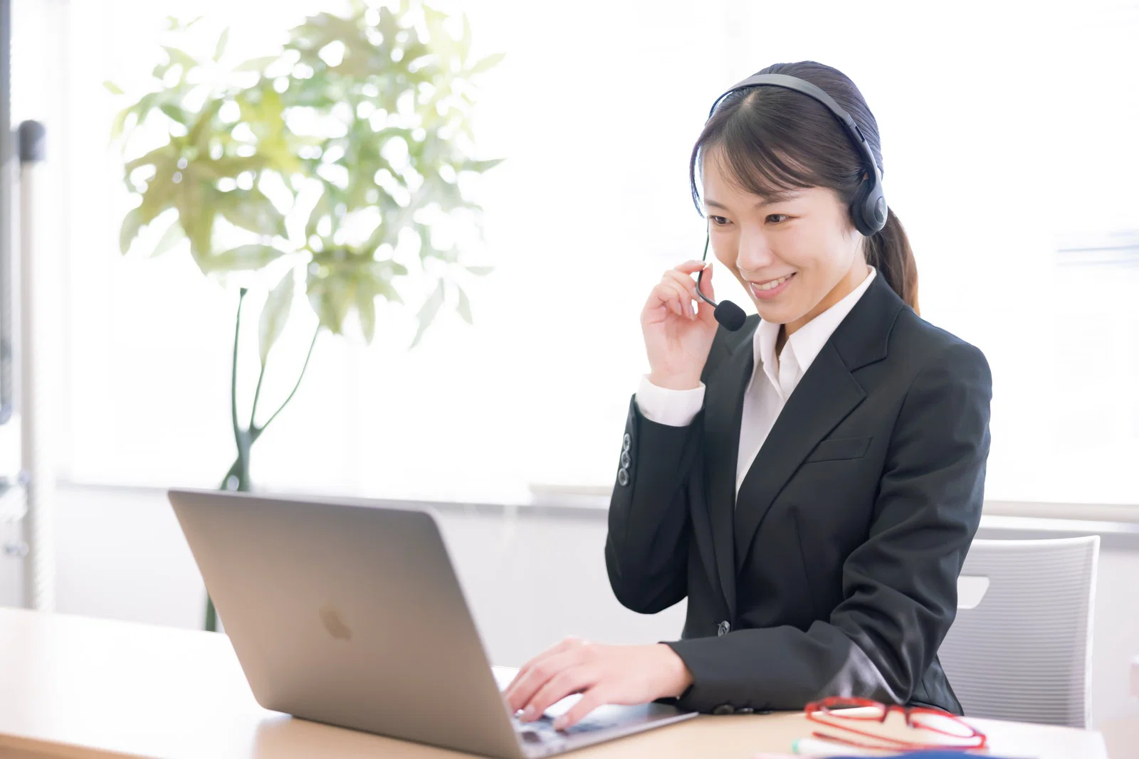 室内でヘッドセットを付けノートPCに向かうスーツ姿の女性社員1人