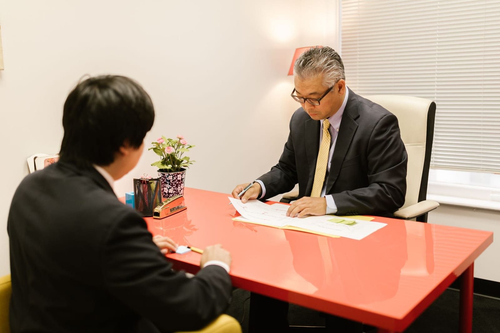 屋内オフィスで赤い机を挟みスーツ姿の男性2人が書類を見ながら向き合う写真
