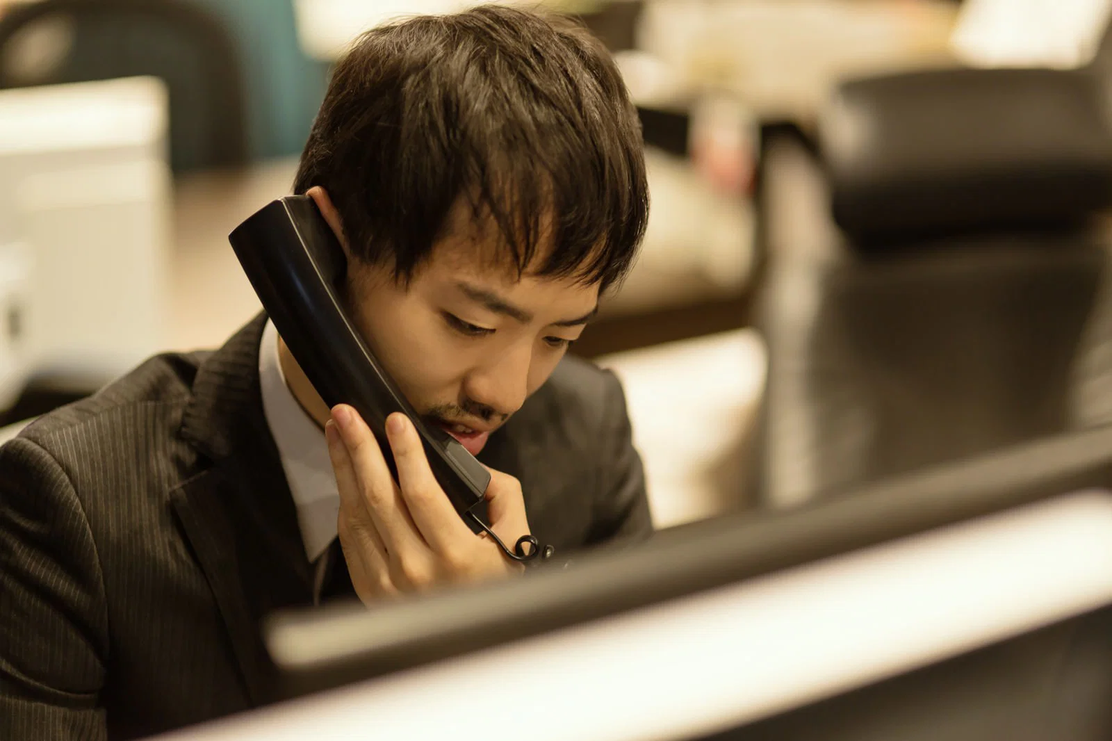 オフィスの自席で受話器を耳に当て眉をひそめる若い男性会社員
