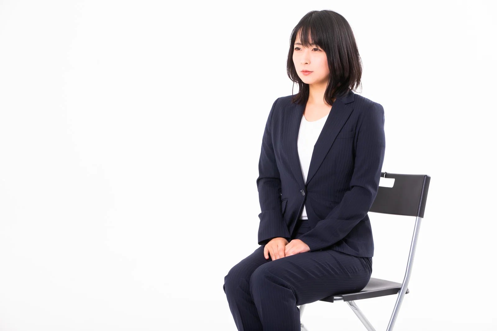 白背景のスタジオで、紺のスーツを着た日本人女性1人がパイプ椅子に座り横を向いている。