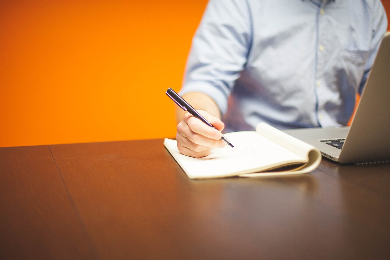 オレンジの壁の前で1人がノートに手書きしパソコンを開いている室内の様子