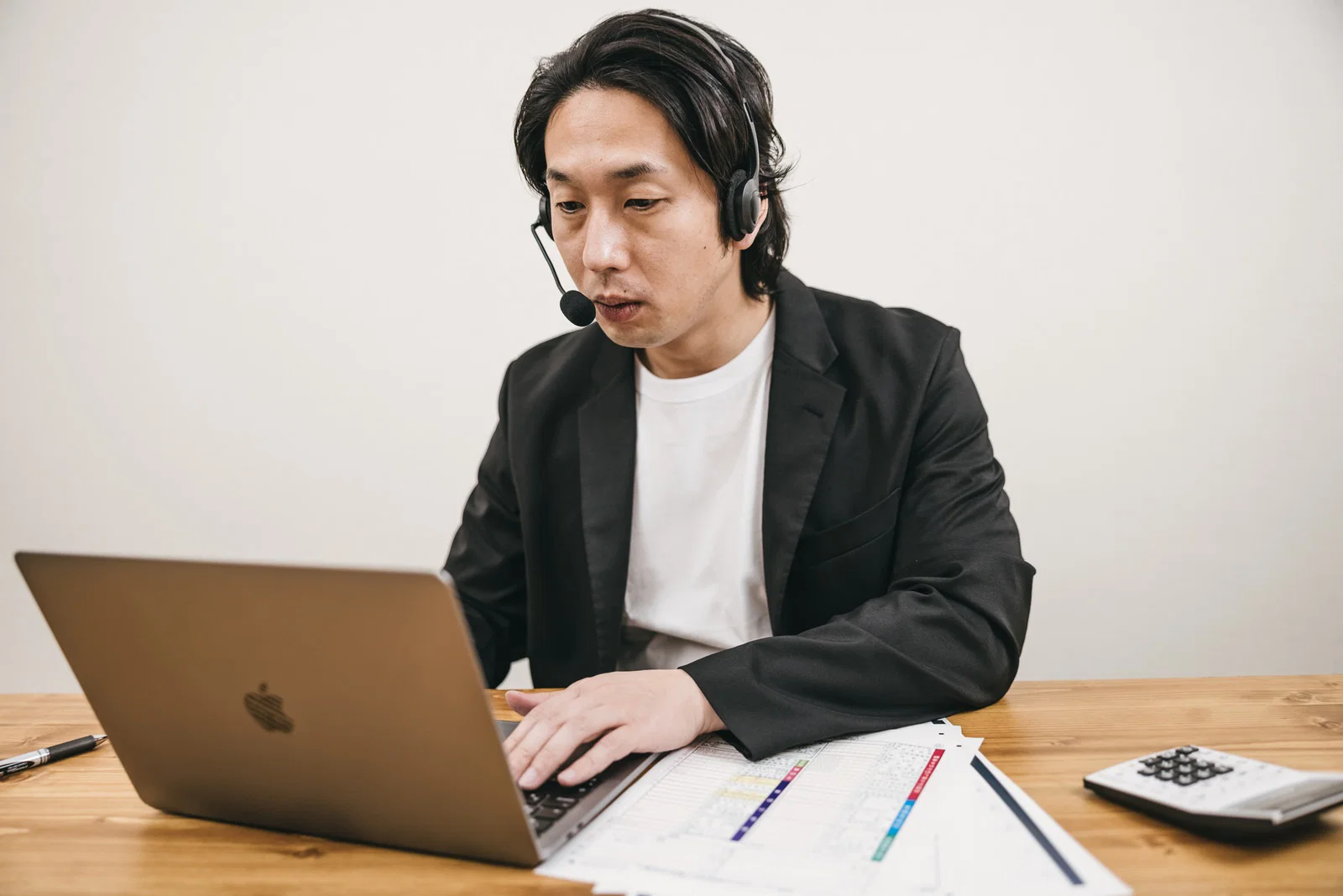 室内で木製デスクに向かい、ヘッドセットを着けてノートPCを操作する男性1人
