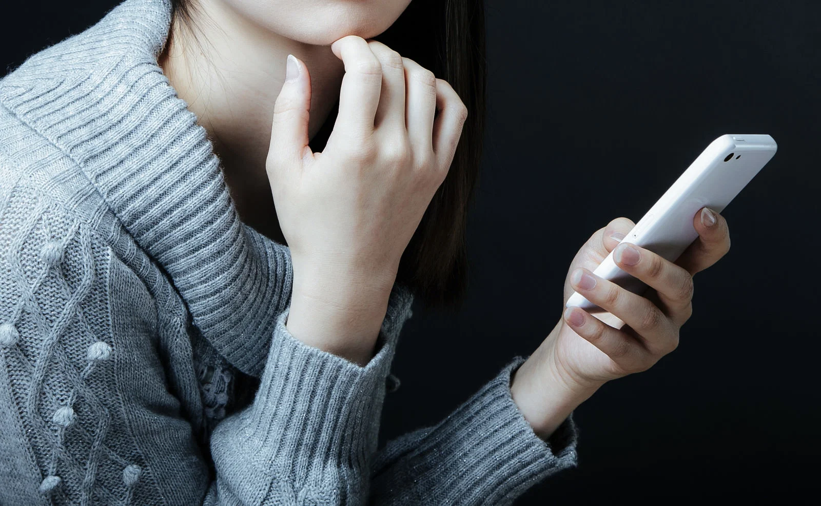 暗い背景の室内でニット姿の女性一人がスマートフォンを手に持ち考え込む様子