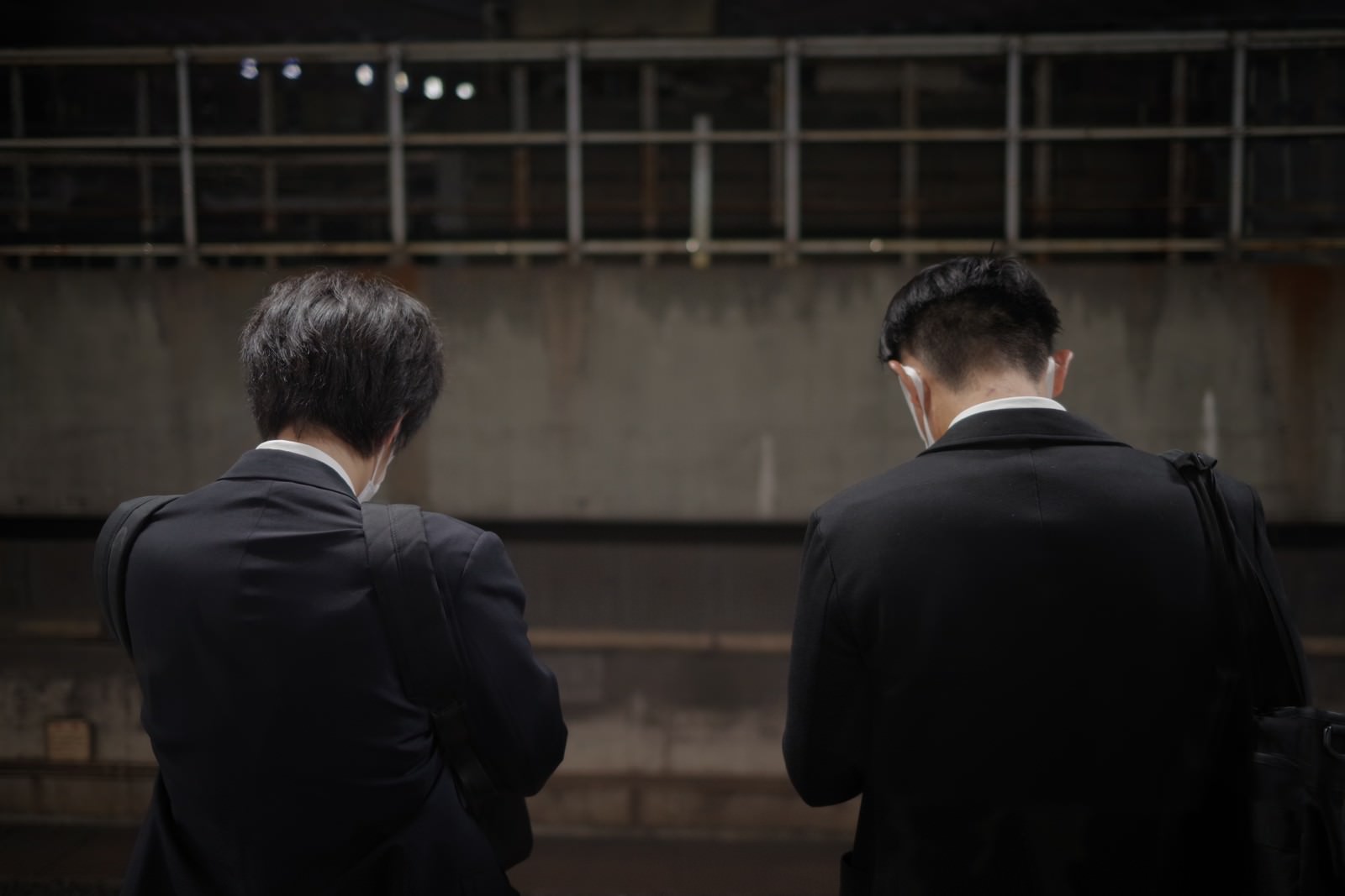 スーツ姿でバッグを肩に掛けた男性2人を背後から撮った写真。2人ともうつむき加減で、背景は薄暗いコンクリート壁と金属柵