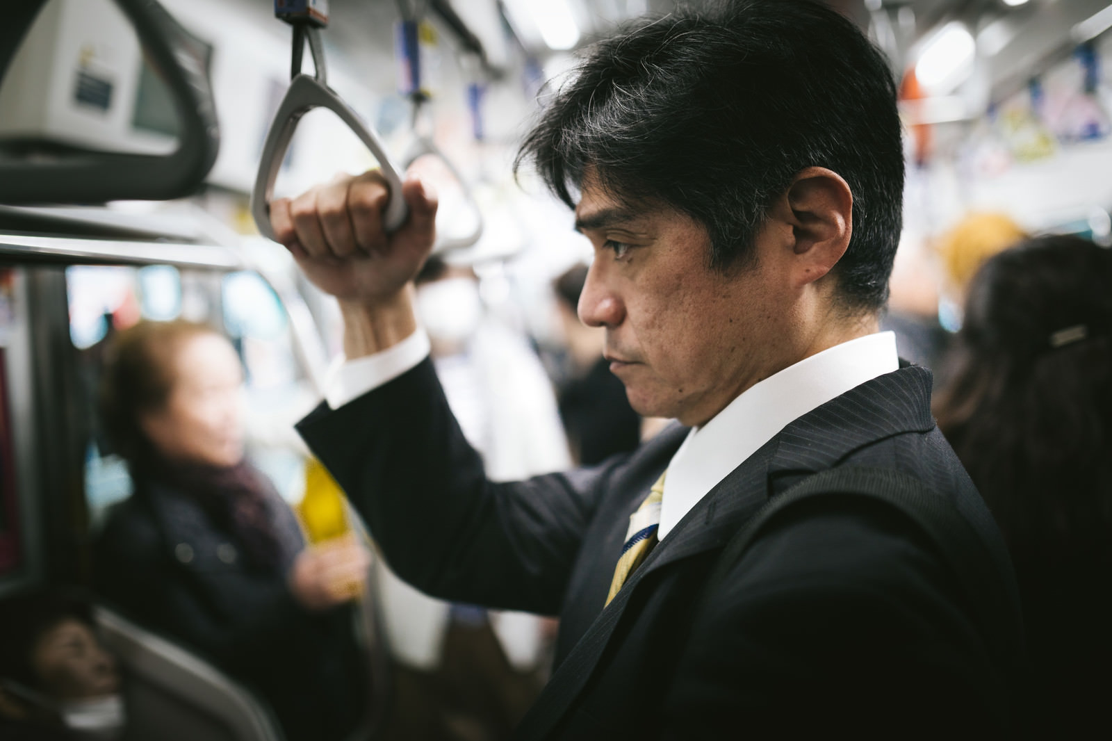 電車内で吊り革をつかんで立つ、スーツにネクタイ姿の中年男性の横顔。背景には他の乗客がぼかして写っている