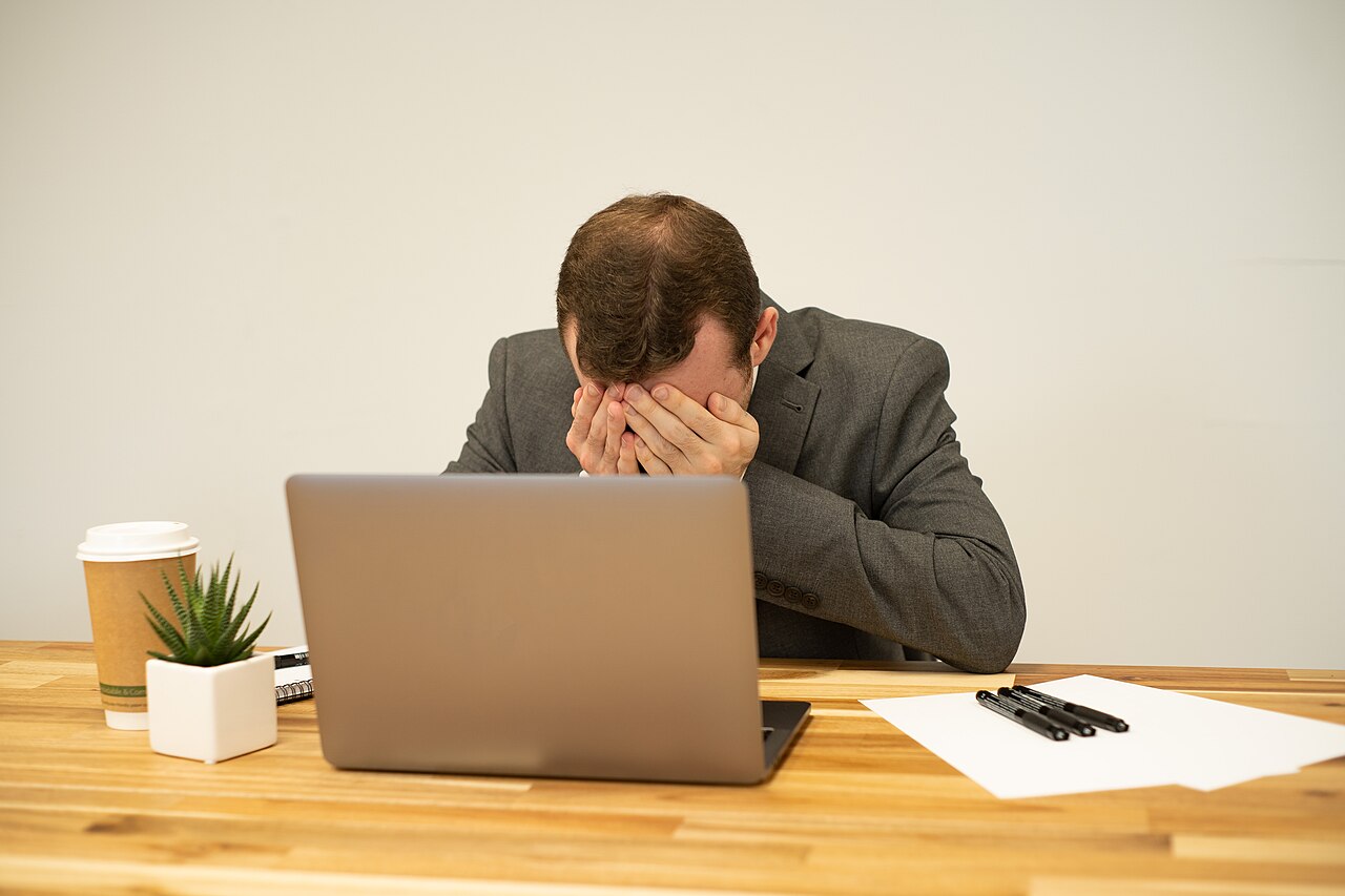 デスクでノートPCの前に座り両手で顔を覆う男性1人、屋内オフィス