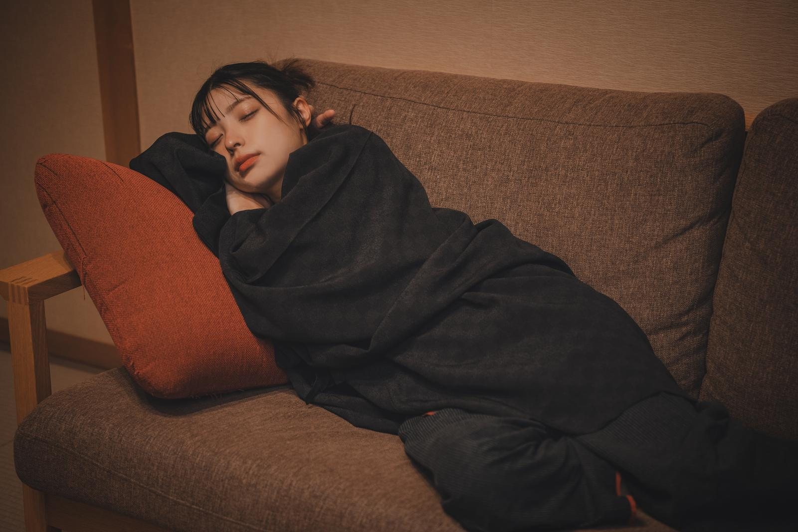 室内のソファで黒い毛布にくるまり眠る若い女性ひとり