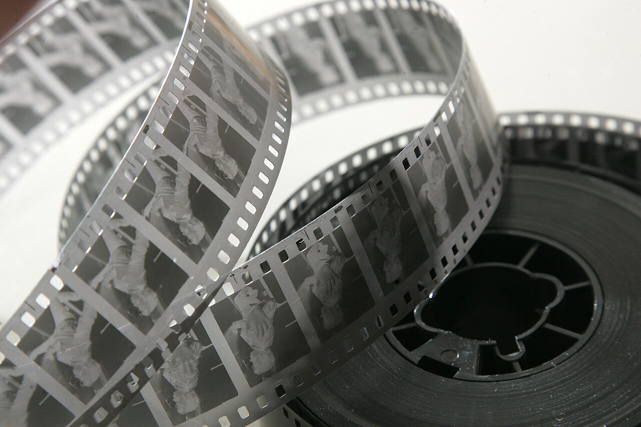 渦を巻いた35mmフィルムのモノクロ写真。送り穴と連続したコマが写り、右側に黒いリールが置かれている
