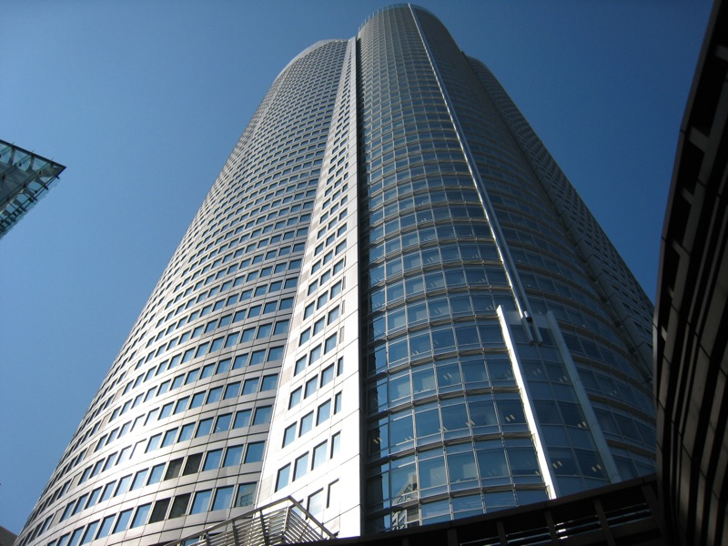 青空の下、真下から見上げた六本木ヒルズ森タワー。金属パネルと角窓の面と曲面のガラス壁が縦に分かれた超高層ビル