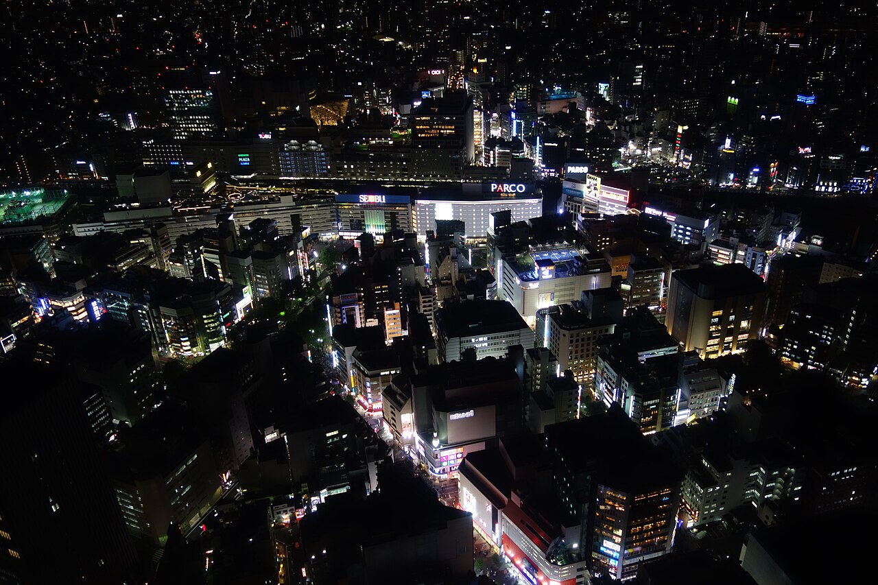 高所から見下ろした夜の池袋の街並み。PARCO、SEIBU、PLAZAなどの看板を掲げた商業ビル群と雑居ビルの窓明かりが広がる