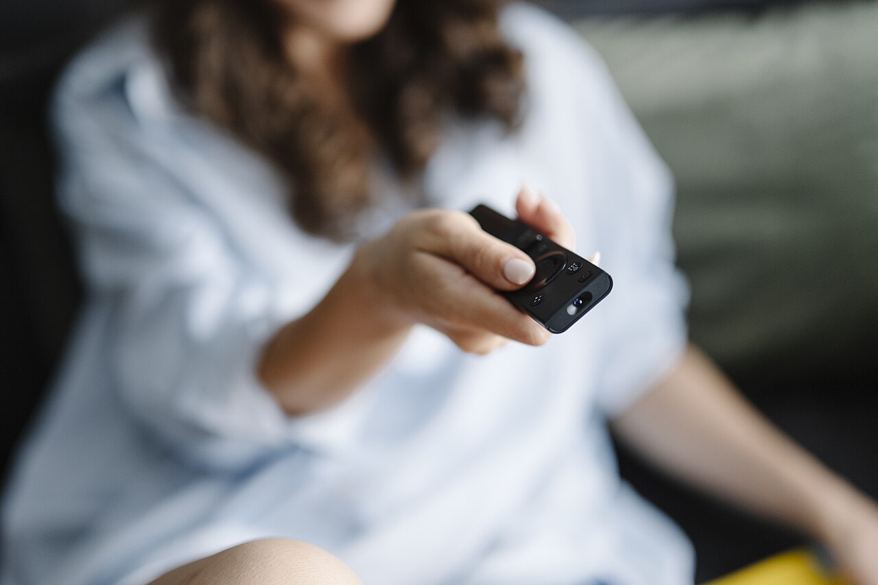 屋内のソファで1人の女性がテレビのリモコンを手に持ち構えている様子