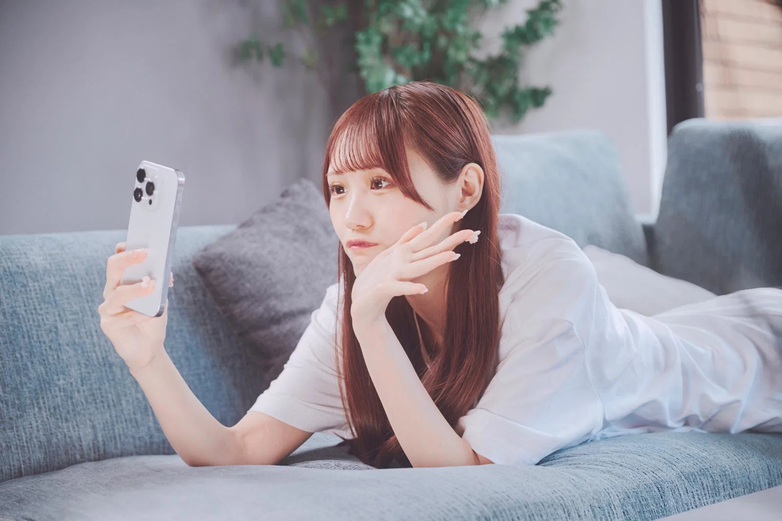 屋内のソファにうつ伏せで寝転びスマホを見る女性1人の写真