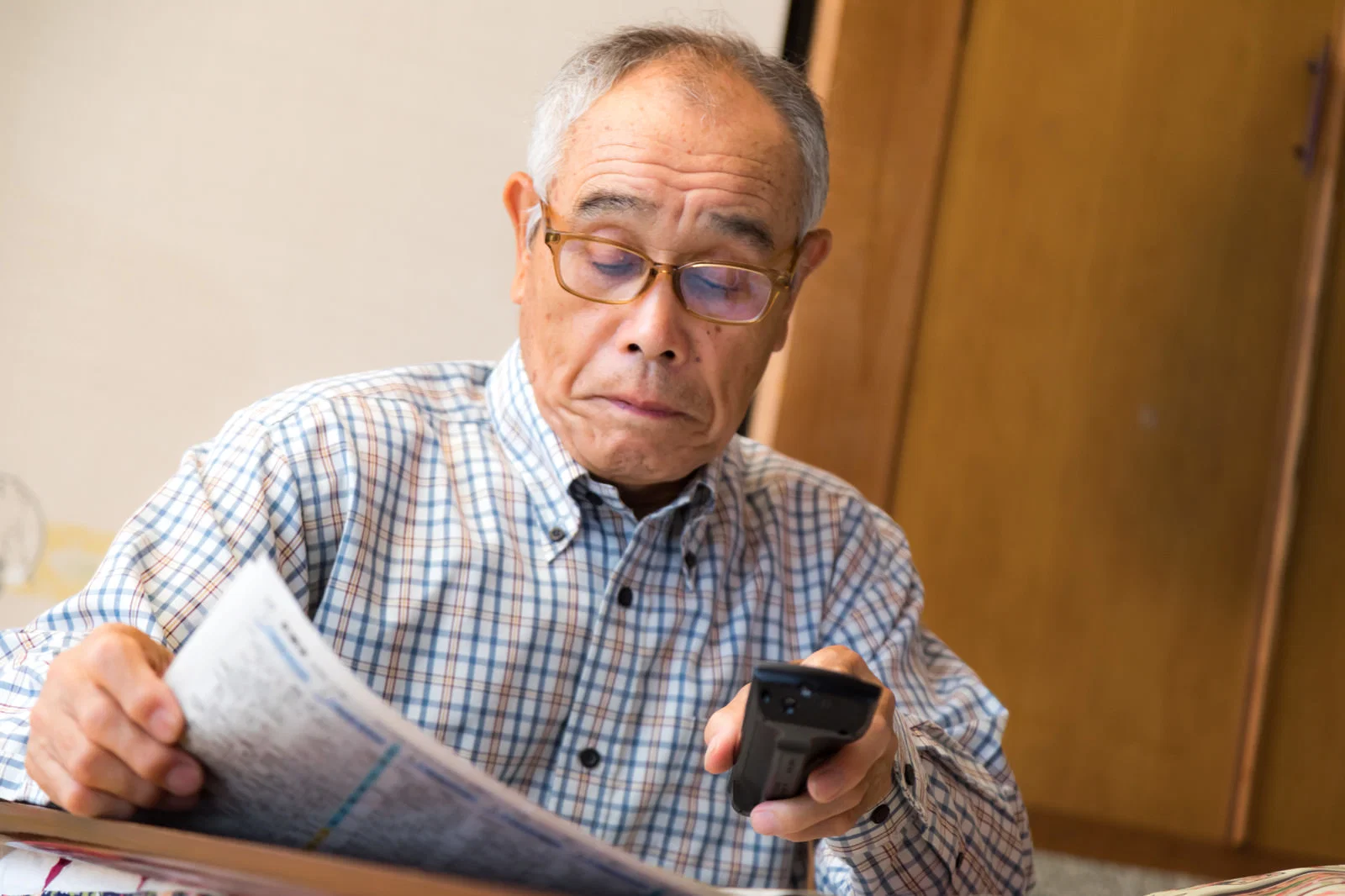 室内で新聞を読みながらリモコンを手に持つ高齢男性1人