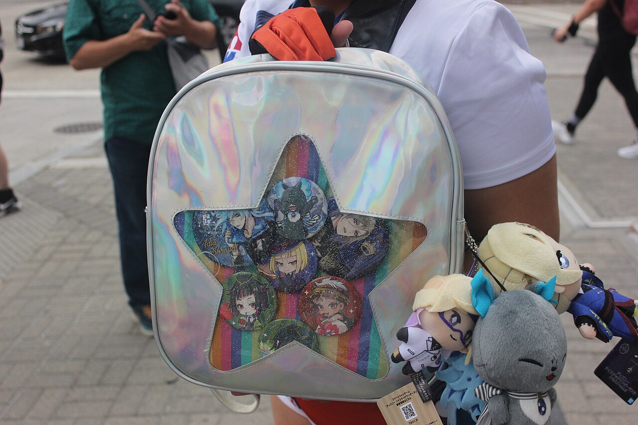 ホログラム調の白いリュックの星形の透明窓にキャラクターの缶バッジが並び、サイドにぬいぐるみマスコットを下げた痛バッグ