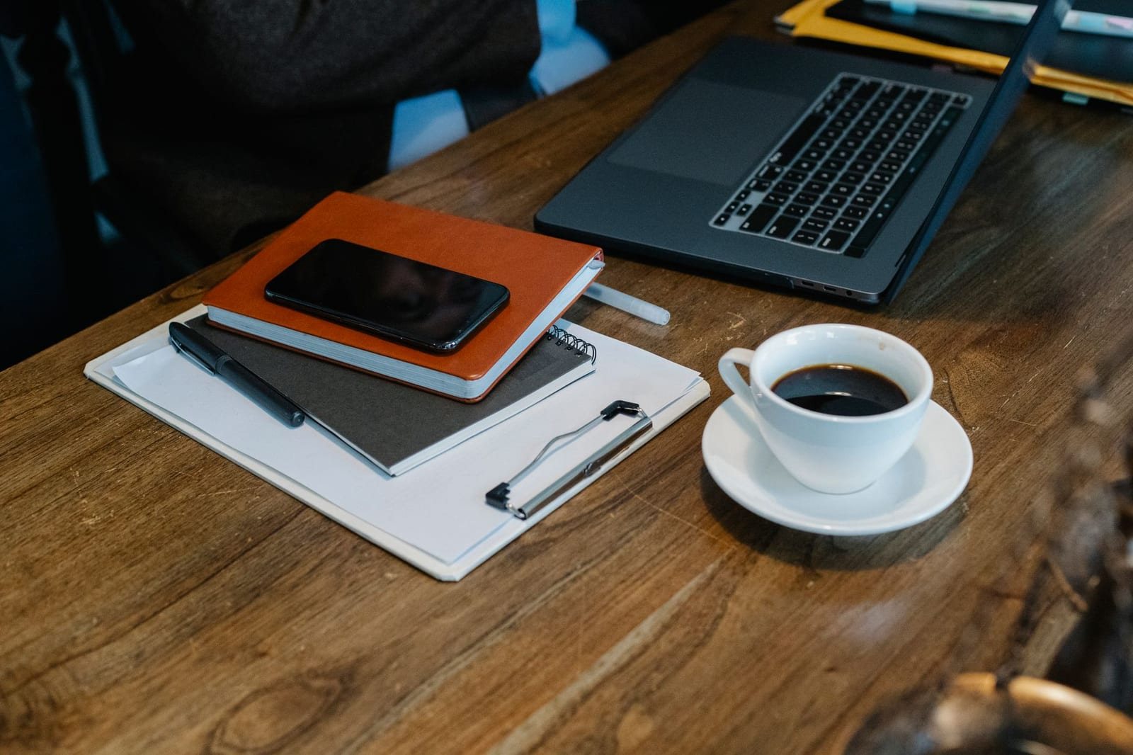 木製テーブルにノートパソコンとノート、スマートフォン、コーヒーカップが置かれた様子