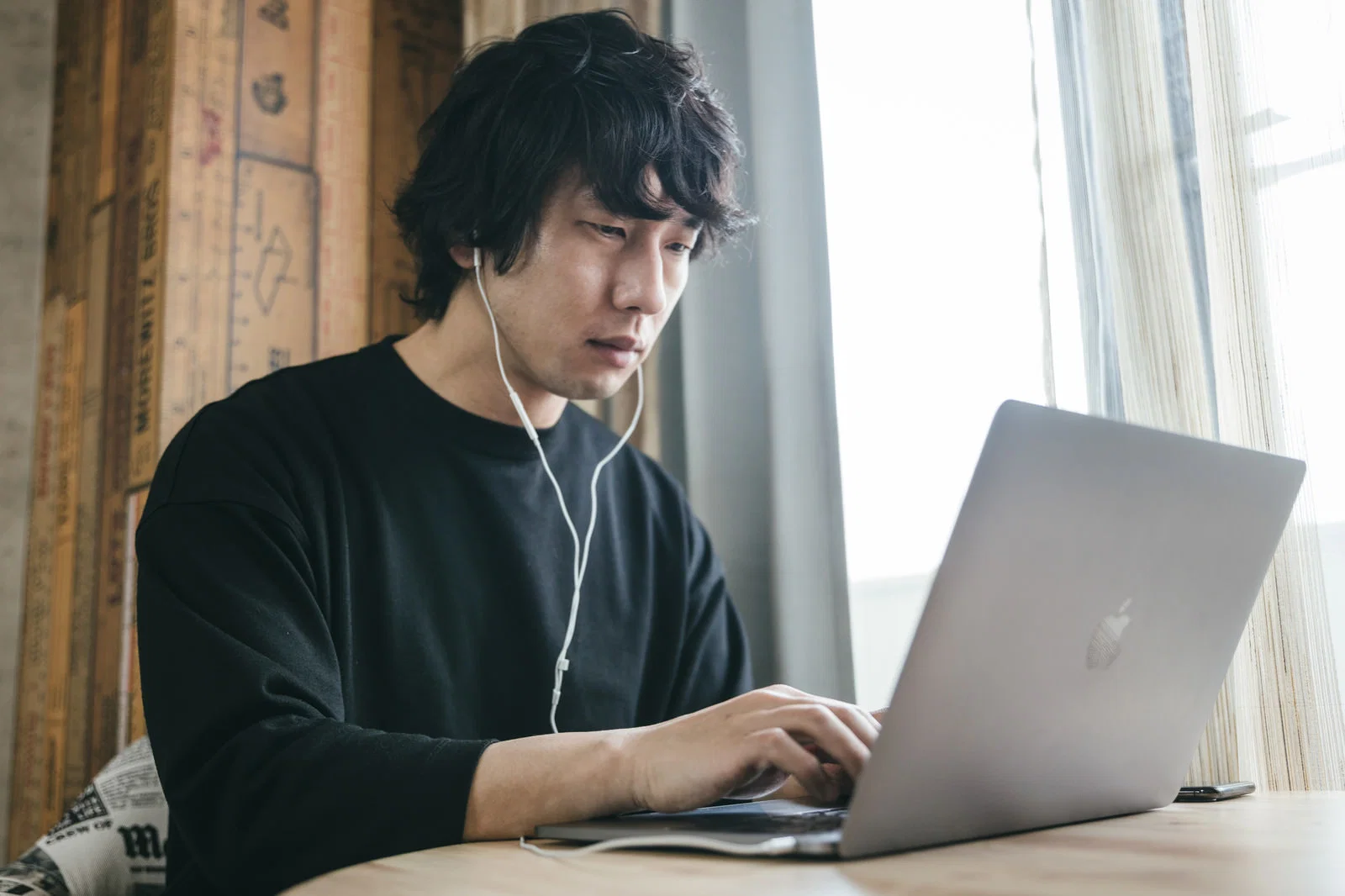 室内の窓際で、イヤホンをつけた男性がノートパソコンを操作している写真。
