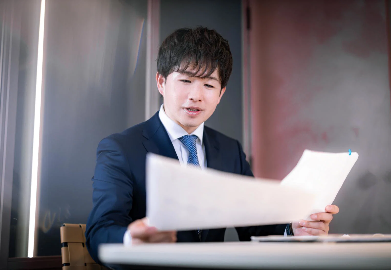 屋内でスーツ姿の男性一人が両手で書類を広げて読んでいる
