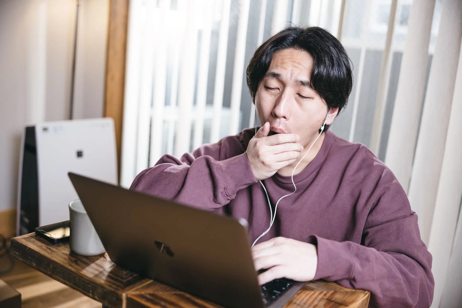 室内でノートパソコンに向かいながら片手で口元を覆いあくびをする男性