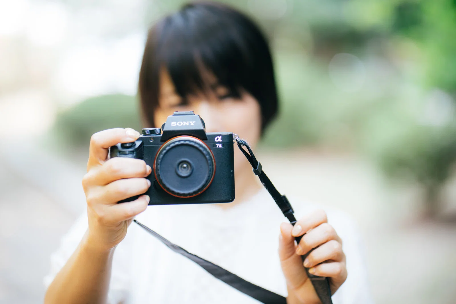 屋外で女性が両手でカメラを構えてレンズをこちらに向けている一枚。