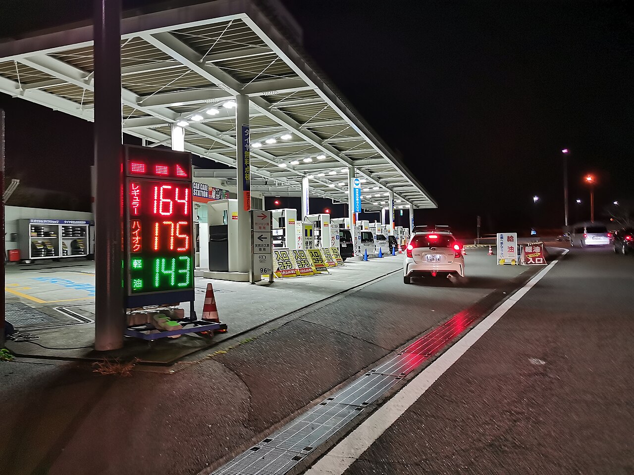 夜間の屋外ガソリンスタンドで、給油する車と料金表示板が並ぶ様子
