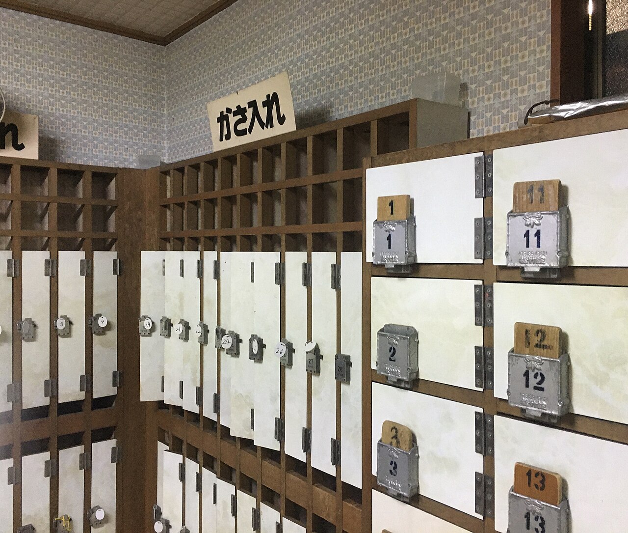 施設の玄関にある木製の集合下足箱。白い扉に番号札付きの金具錠が並び、上部の格子棚には「かさ入れ」と書かれた貼り紙がある