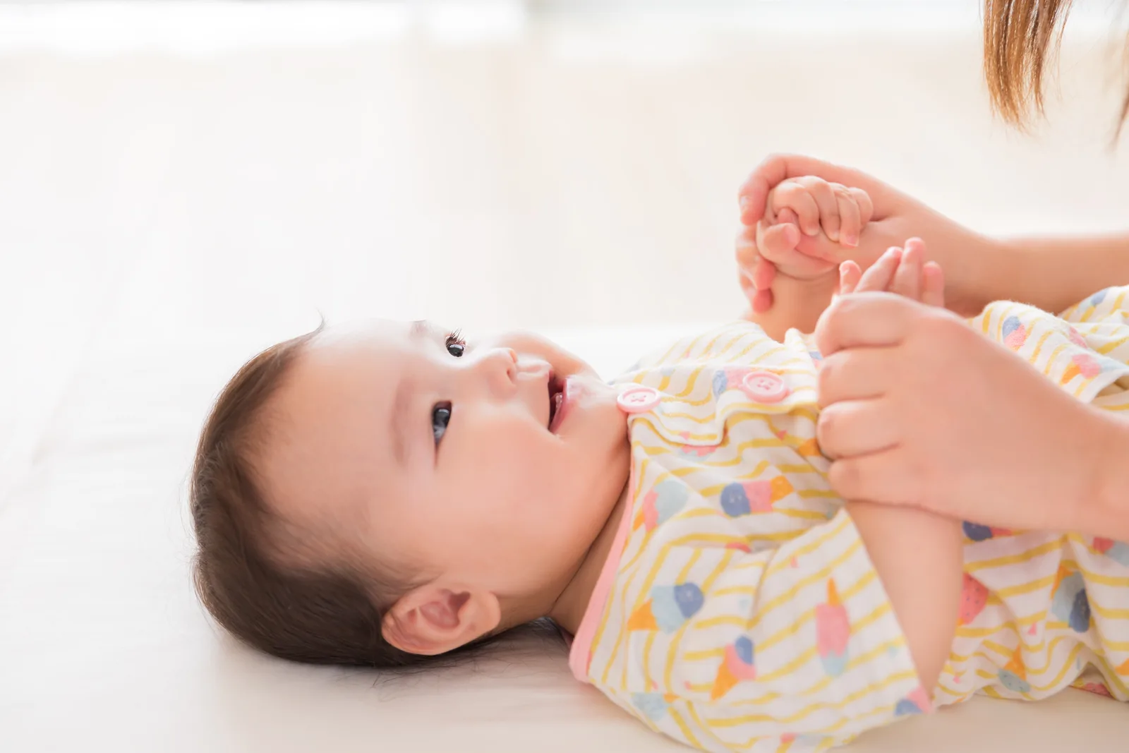 白い布の上で仰向けに笑う赤ちゃんの両手を、大人の手が優しく握っている
