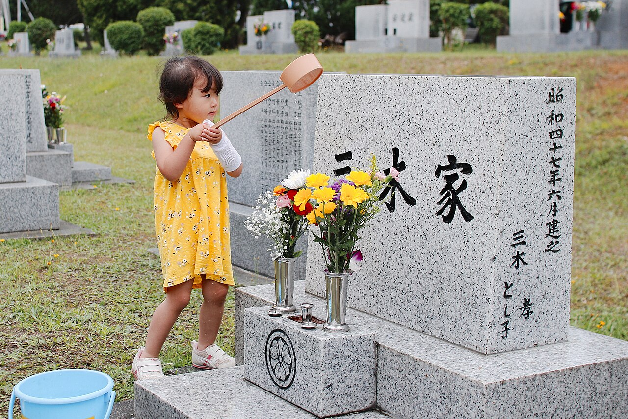 屋外の墓地で黄色いワンピースの女児が柄杓で墓石に水をかけている一人の写真