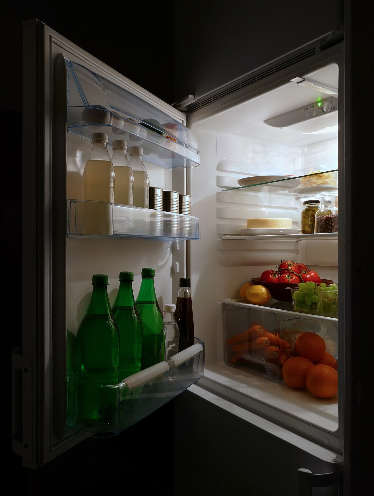 扉を開けた冷蔵庫の庫内。扉ポケットに瓶や飲料が並び、棚にはチーズや瓶詰め、トマト・ぶどう・レモン、野菜室にニンジンとオレンジが入っている