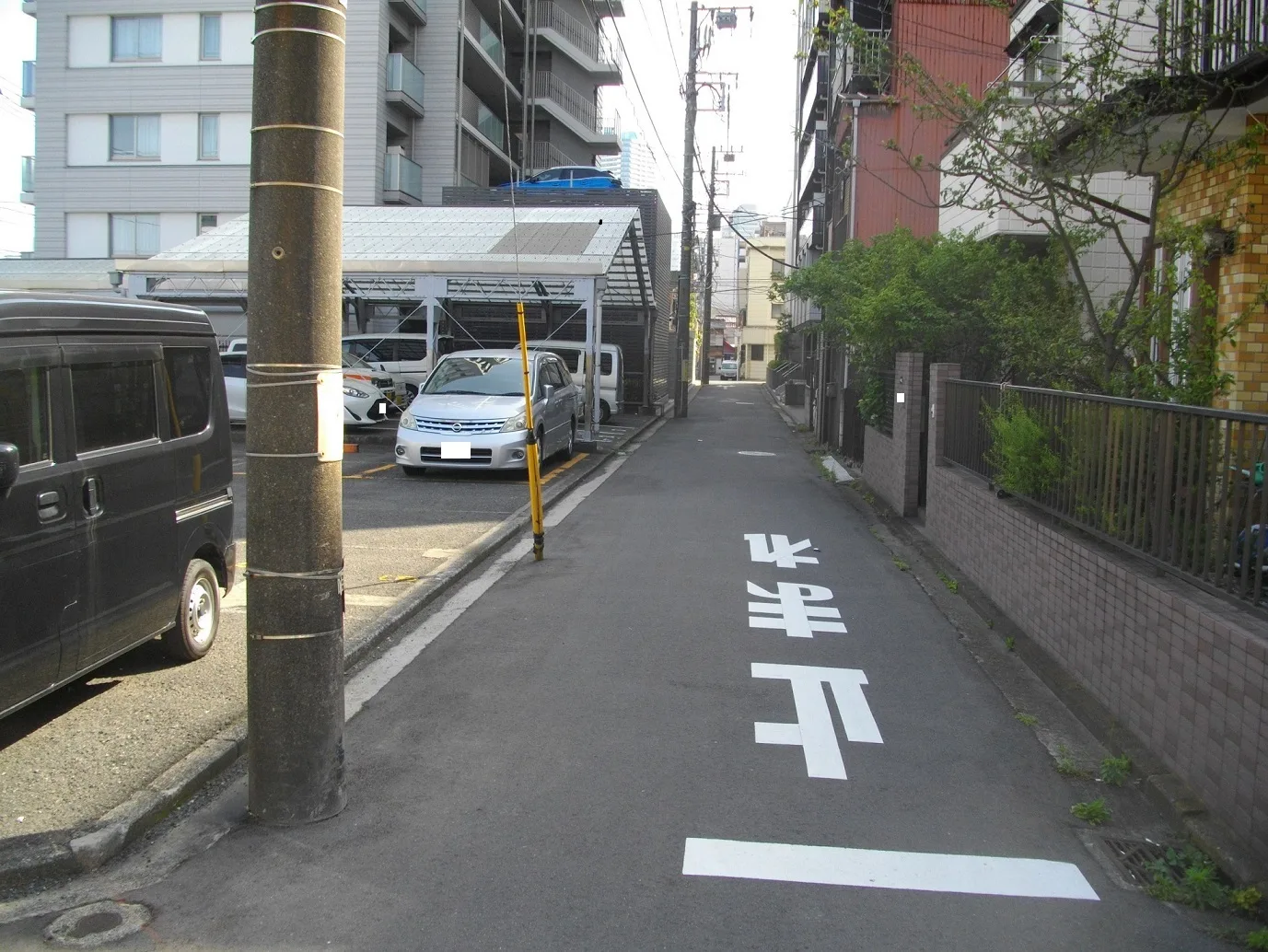 住宅地の中央線がない生活道路。手前の路面に白い「止まれ」の文字と停止線、左側に電柱と月極駐車場、右側にブロック塀と住宅が続く