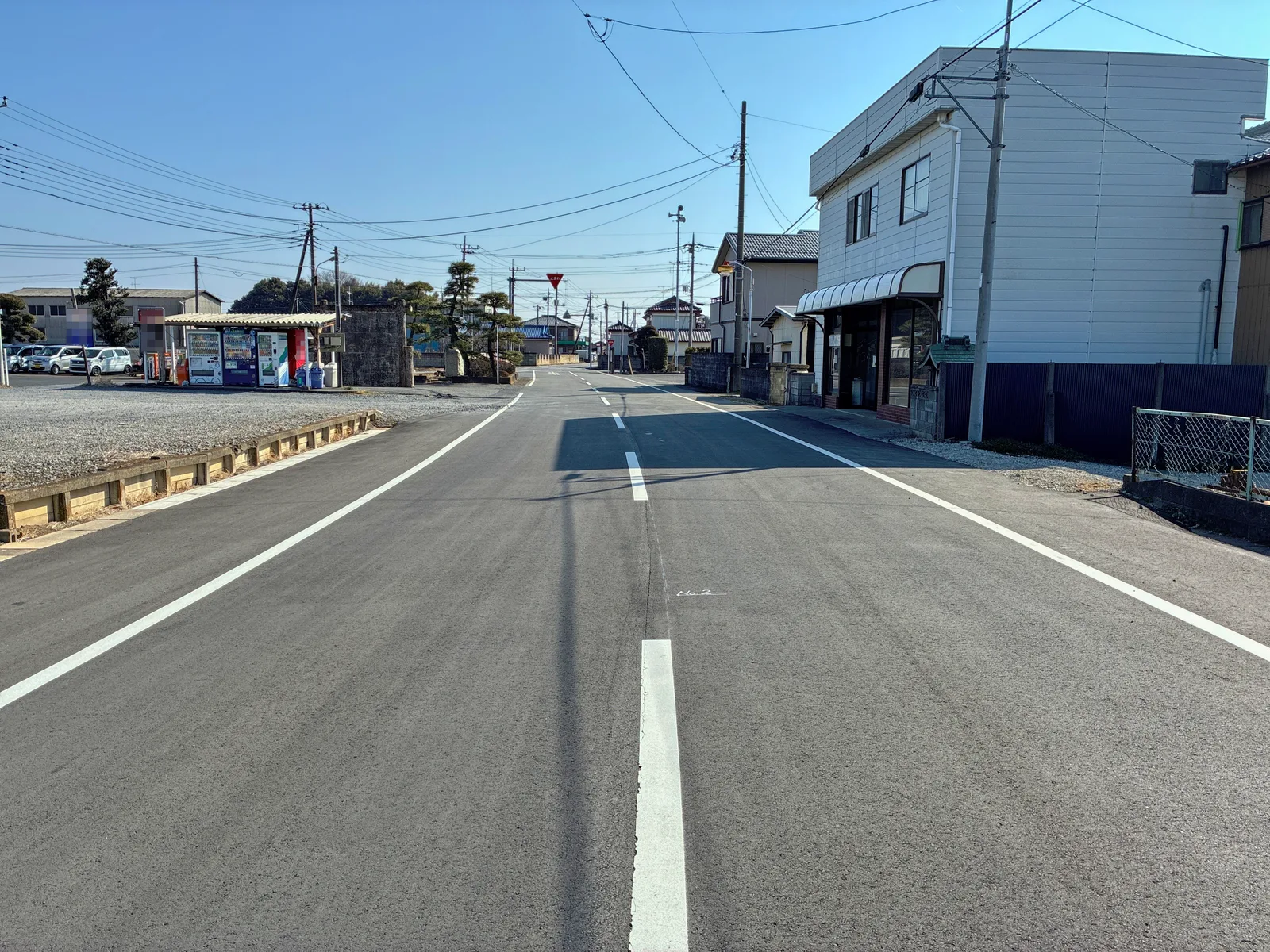 郊外の2車線道路。白い外側線と白の破線の中央線が引かれ、左に自動販売機と駐車場、右に住宅、奥に赤い止まれ標識が見える