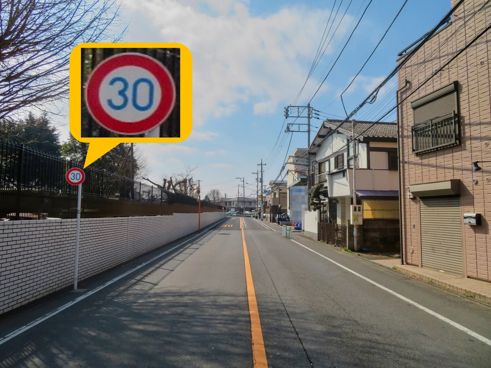 市街地の直線道路。左側の白い柵沿いに最高速度30km/hの赤丸標識が立ち、吹き出しで拡大表示されている。車道中央にはオレンジの中央線があり路面にも30の表示