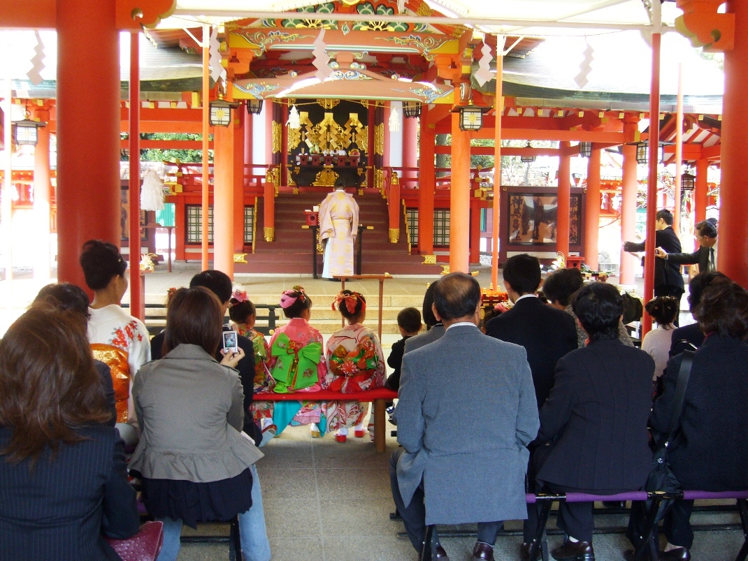 朱色の神社拝殿で晴れ着の子どもと大勢の参列者が祈祷を受けている