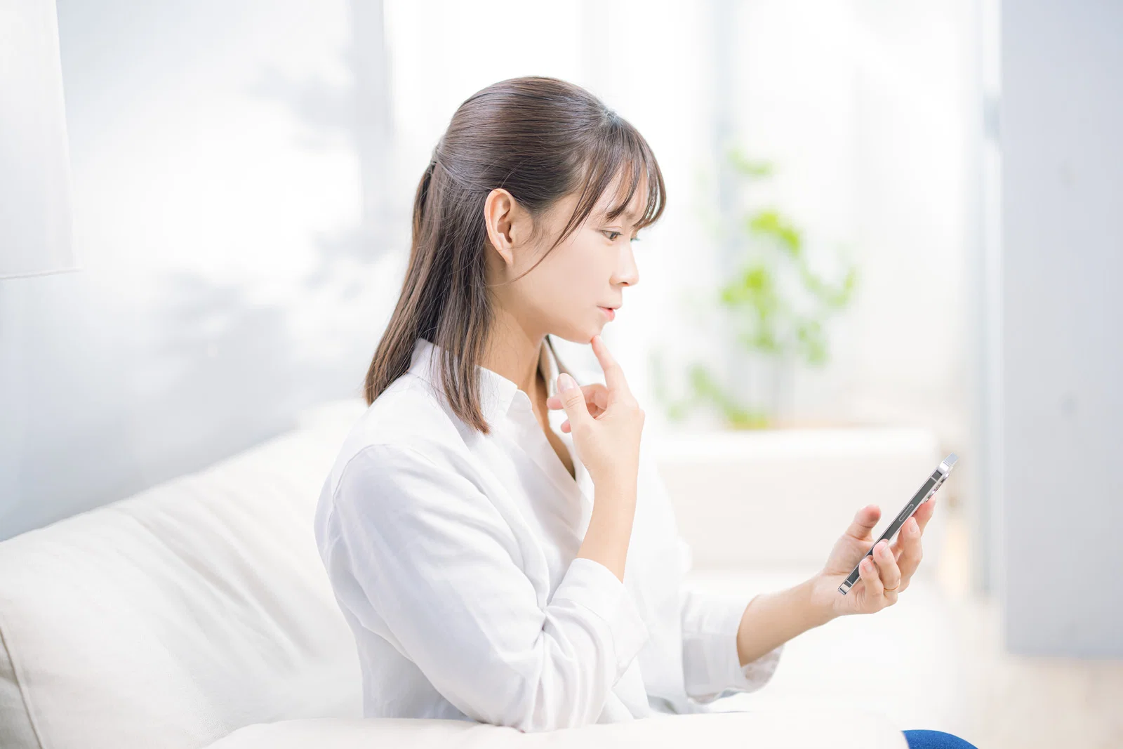 室内のソファで白いシャツの女性1人がスマートフォンの画面を見つめている
