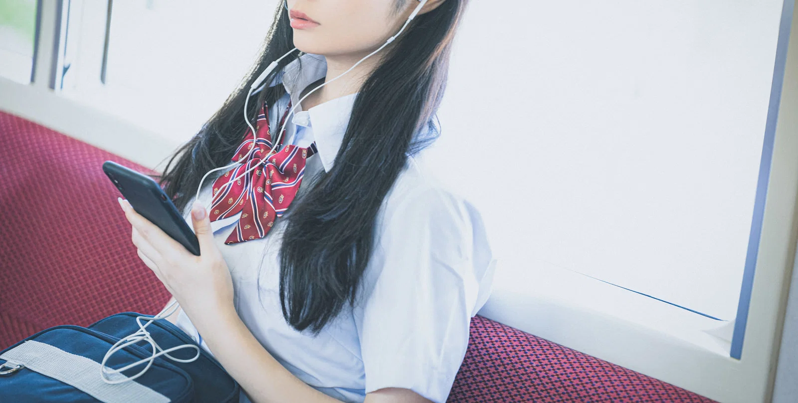 電車の座席でイヤホンをつけスマートフォンを見る女子学生