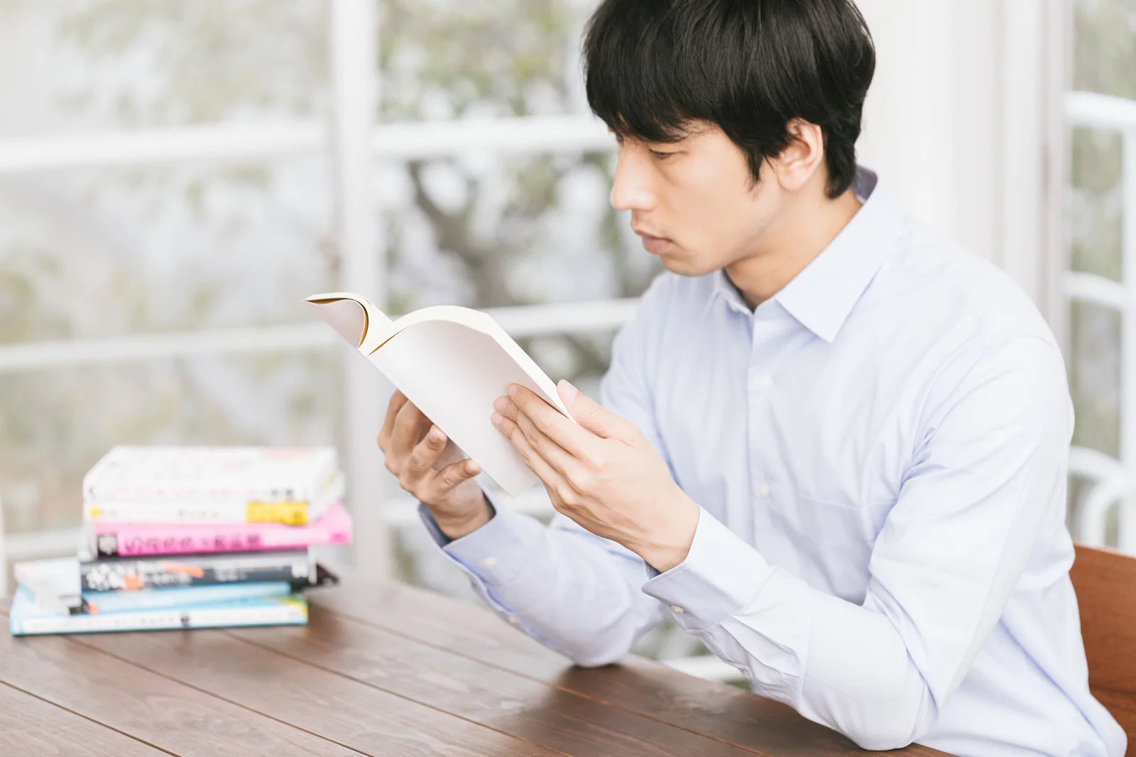屋内の木製テーブルで1人の男性が本の山を横に読書している様子