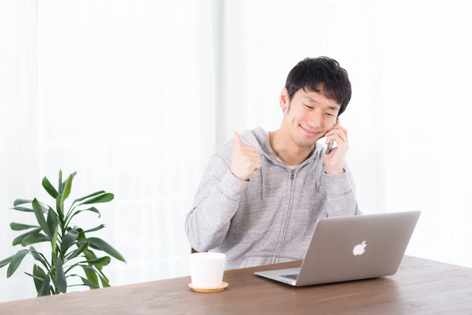 自宅の机でノートパソコンを前に電話しながら親指を立てる男性
