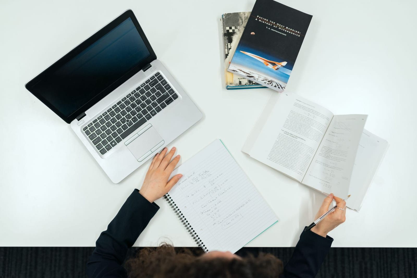 白い机にノートPCと本、ノートを広げ人物1人が勉強している写真