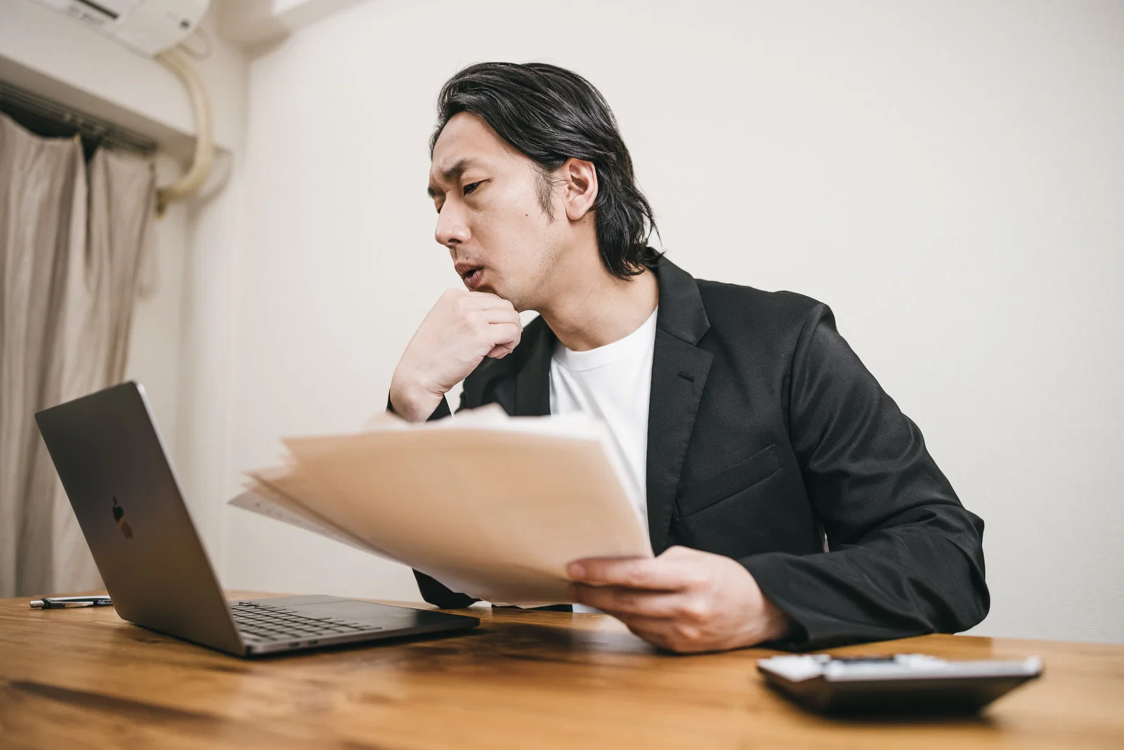 室内でスーツ姿の男性がノートPCの前で書類を手に考え込む様子
