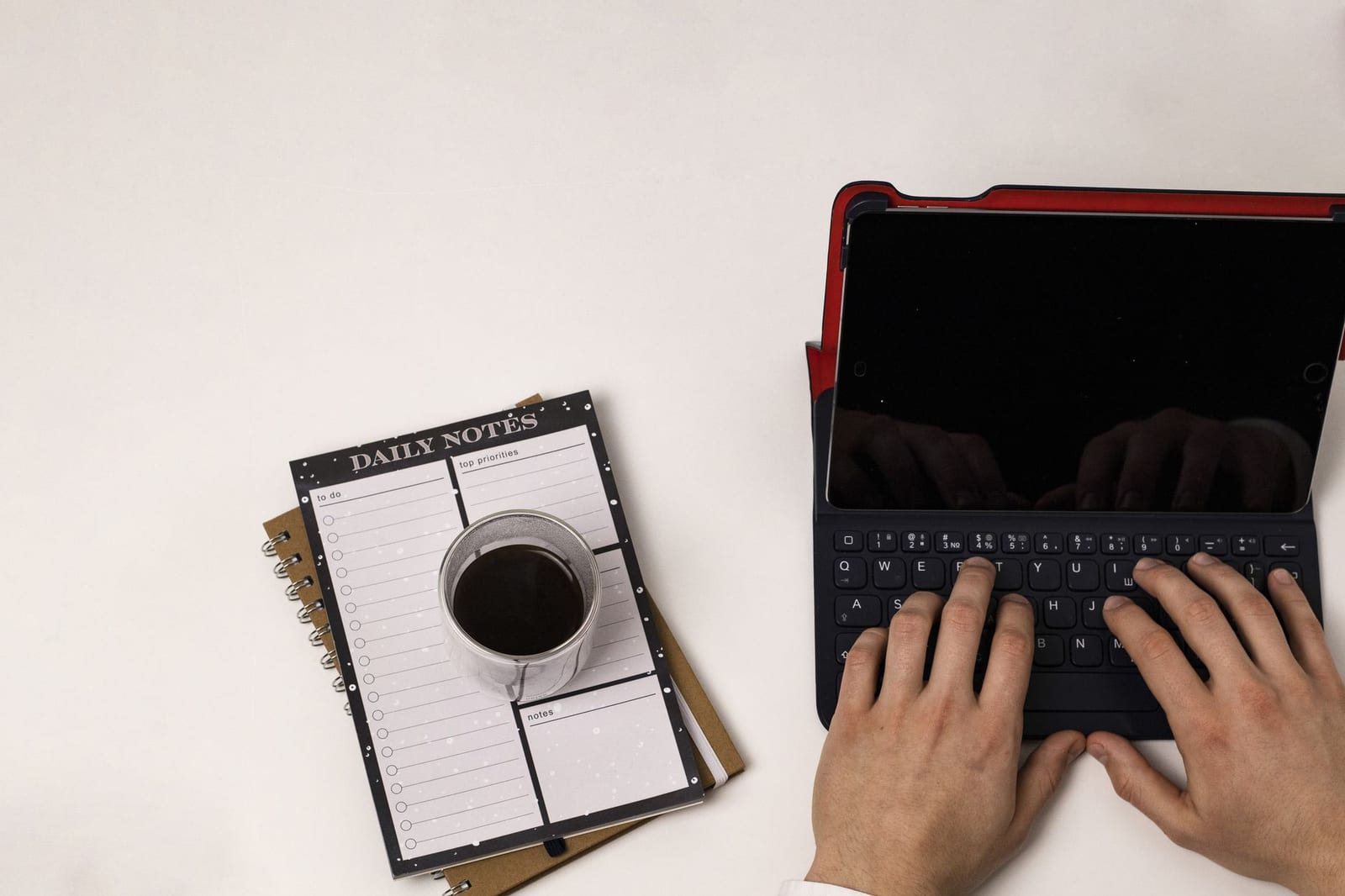 白い机の上でノートとコーヒーの横、タブレット付きキーボードを両手で操作する俯瞰写真。