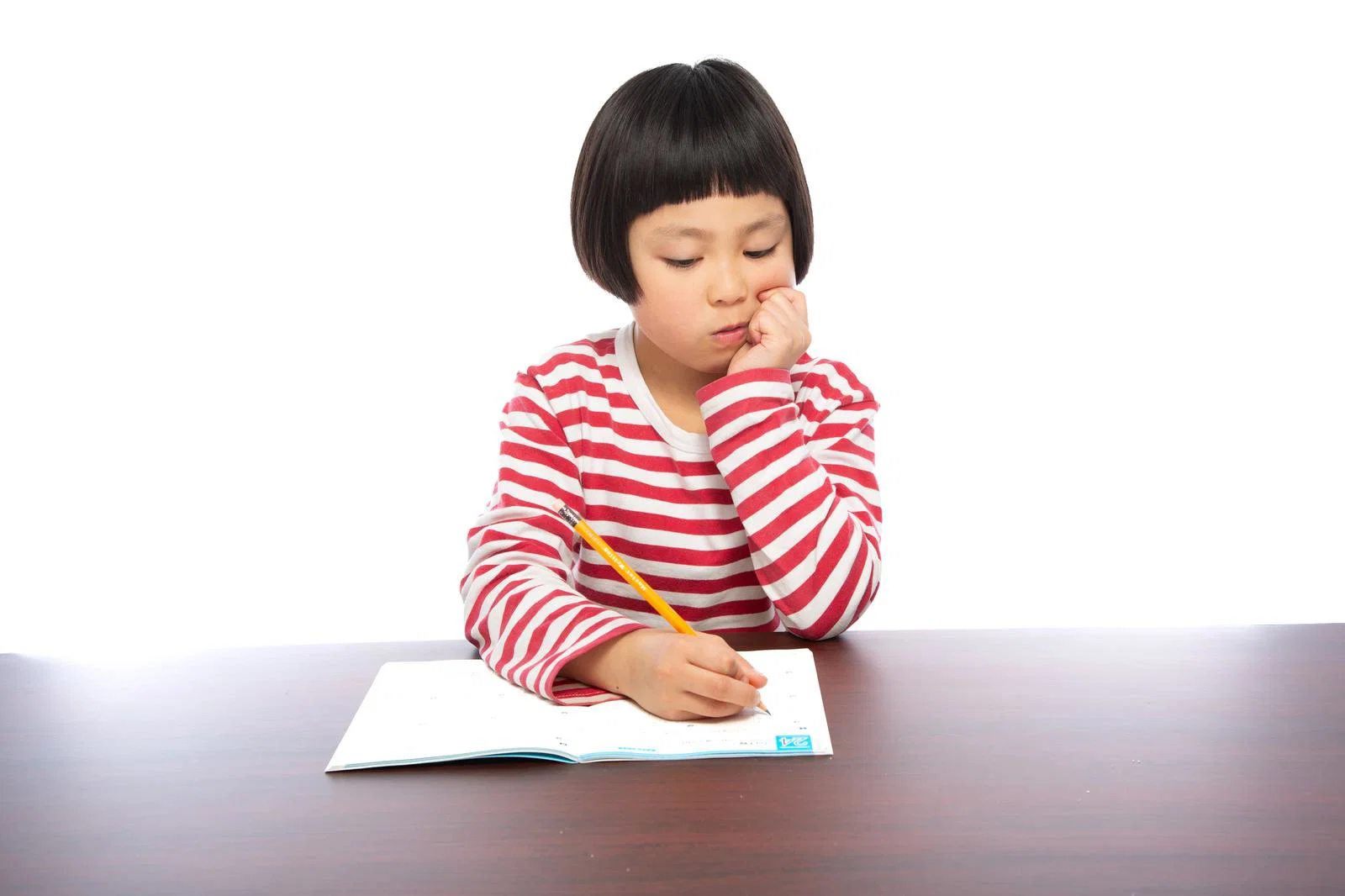白背景の室内で縞模様の服の女の子1人が鉛筆を持ち机の上のノートに書き込んでいる写真