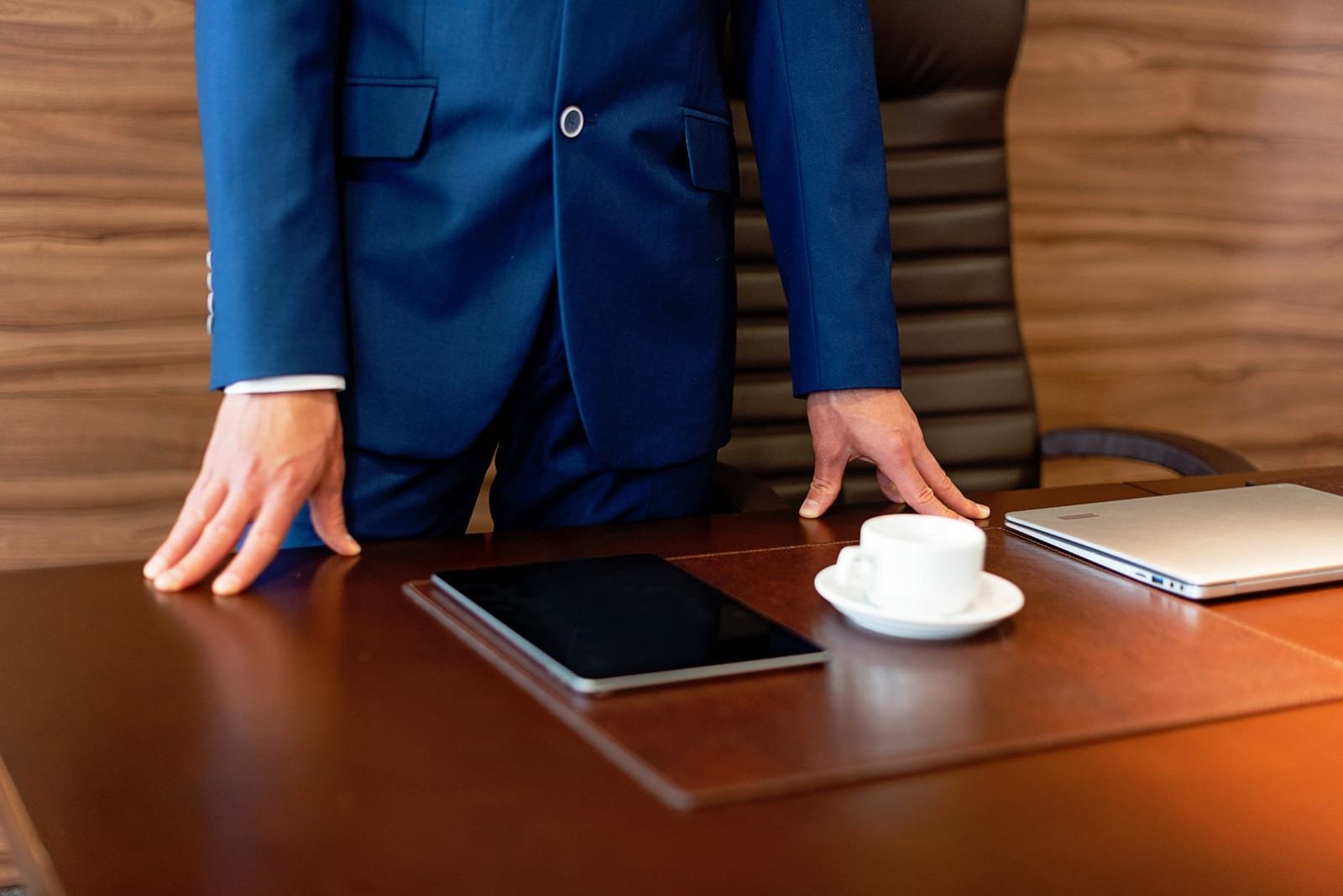 室内の机にタブレットとノートパソコン、コーヒーを置いてスーツ姿の男性が立つ