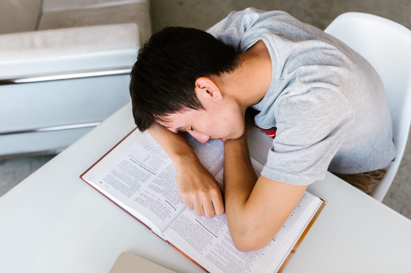屋内の机に置いた本に顔を伏せて眠る若い男性一人の写真